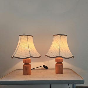 Pu&ograve; includere: Due lampade da tavolo in legno con paralumi in tessuto bianco. I paralumi hanno un bordo a festoni e un bordo marrone. Le lampade sono accese e illuminano la stanza.