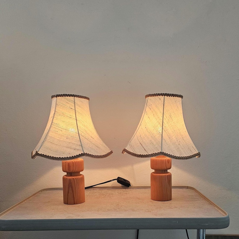 Pu&ograve; includere: Due lampade da tavolo in legno con paralumi in tessuto bianco. I paralumi hanno un bordo a festoni e un bordo marrone. Le lampade sono accese e illuminano la stanza.