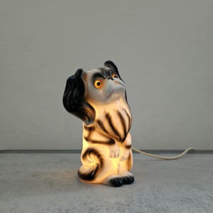 Op de afbeelding: Een keramische hondvormige lamp in zwart en wit. De lamp heeft een nieuwsgierige uitdrukking en gloeiende oranje ogen. Licht schijnt door het lichaam en benadrukt de markeringen en kenmerken van de hond. Een wit snoer loopt vanaf de basis.
