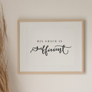 Può includere: Una stampa incorniciata con la scritta "HIS GRACE IS Sufficient" in corsivo nero. La stampa ha un passe-partout bianco e una cornice in legno chiaro. L'immagine è esposta su una parete di colore neutro, con erba ornamentale secca nell'angolo.