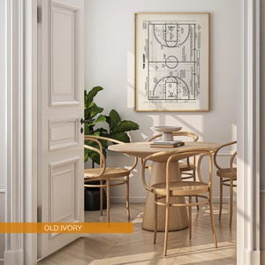 Op de afbeelding: Een ingelijste basketbalveldtekening hangt boven een ronde houten tafel en stoelen. De kamer heeft een neutraal kleurenpalet met een grote plant in de hoek. Het deurkozijn is geschilderd in "Old Ivory".