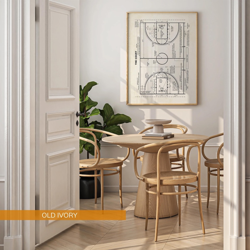 Op de afbeelding: Een ingelijste basketbalveldtekening hangt boven een ronde houten tafel en stoelen. De kamer heeft een neutraal kleurenpalet met een grote plant in de hoek. Het deurkozijn is geschilderd in "Old Ivory".