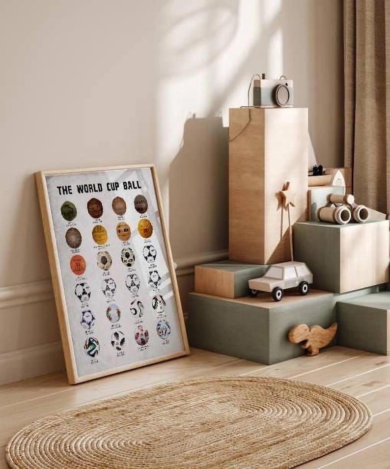 May include: A framed poster titled THE WORLD CUP BALL featuring a grid of various soccer ball illustrations in different colors and patterns, displayed in a light wood frame next to stacked wooden blocks and a small toy car.