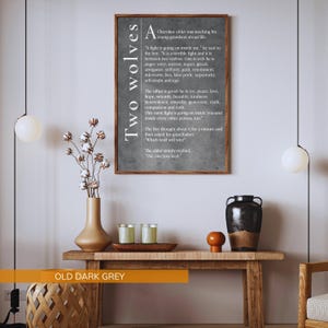 a wooden table with a vase of flowers, a black vase, and a wooden bowl on it. Above the table, there is a framed text that reads "Two wolves" and a quote underneath it. The table is set against a white wall, and there are two hanging lights above it.