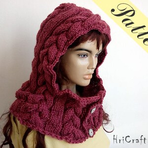 May include: A burgundy knitted hooded cowl with a cable knit pattern and two buttons. The cowl is worn by a person with long brown hair.