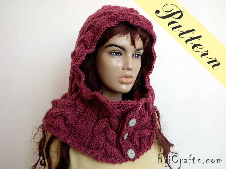 May include: A burgundy knitted cowl with a hood and button closures. The cowl is made of a soft, chunky yarn and has a cable knit pattern.
