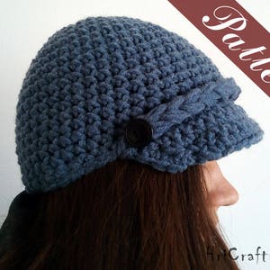 May include: A blue crocheted hat with a brim and a button detail. The hat is worn by a person with long brown hair.