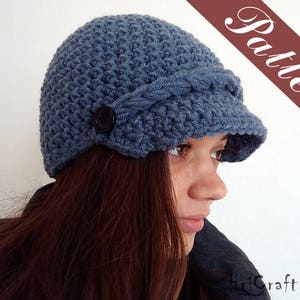 May include: A blue crocheted newsboy hat with a braided detail and a black button. The hat has a wide brim and is worn by a person with long brown hair.