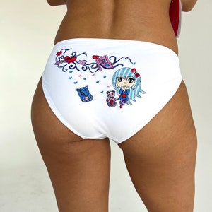以下が含まれることがあります： 青い髪の女の子と2匹のぬいぐるみクマが描かれたカラフルな漫画のデザインが施された白い綿の下着。デザインにはハート、渦巻き、花が含まれています。