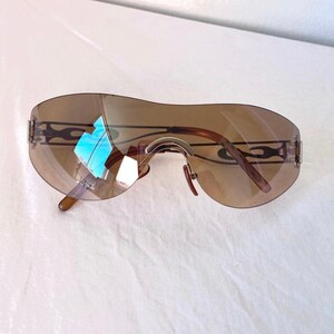 May include: Brown rimless sunglasses with a curved design. The lenses are a gradient brown color. The temples have a decorative black flame design. The sunglasses have a silver bridge and nose pads.