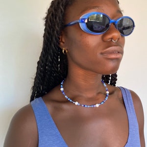 May include: A blue beaded necklace with white pearls and blue beads. The necklace is worn with blue oval sunglasses and a blue tank top. The sunglasses have a blue frame and dark lenses. The model is wearing gold hoop earrings.