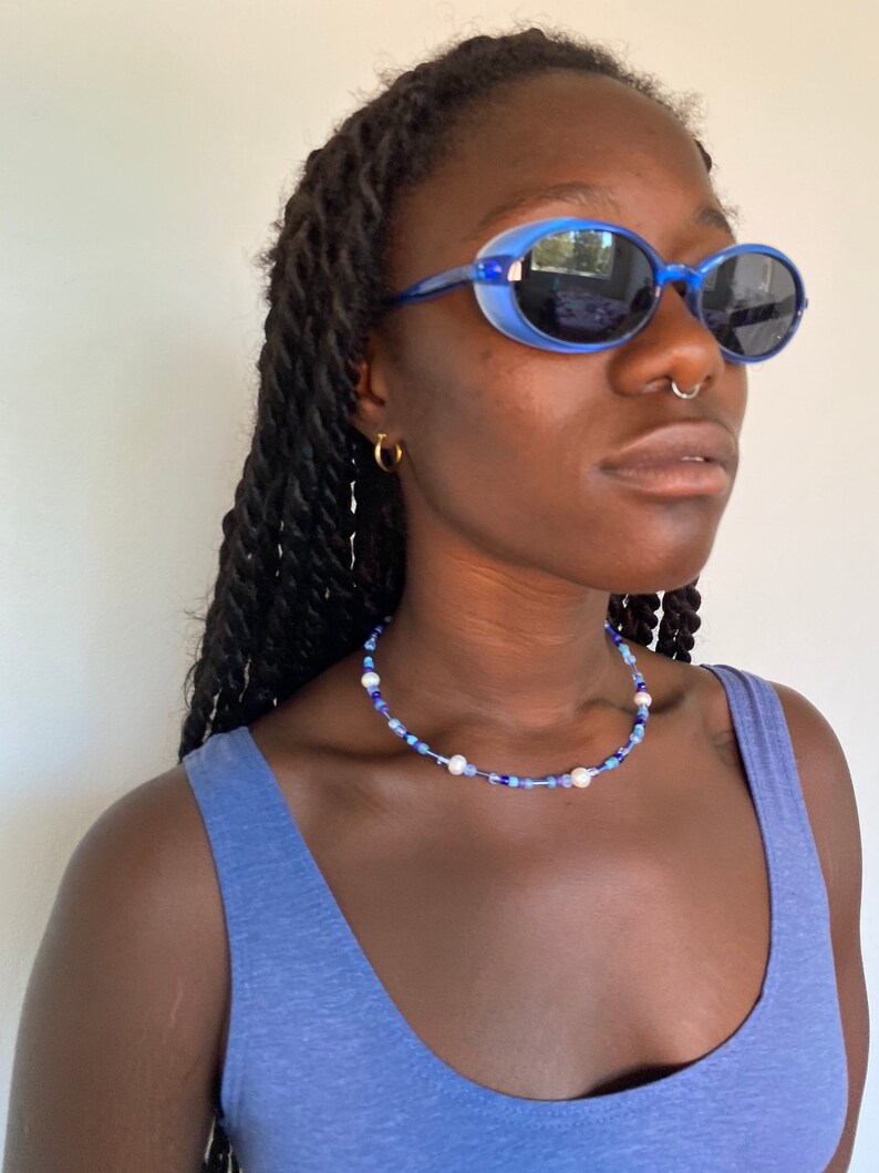 May include: A blue beaded necklace with white pearls and blue beads. The necklace is worn with blue oval sunglasses and a blue tank top. The sunglasses have a blue frame and dark lenses. The model is wearing gold hoop earrings.