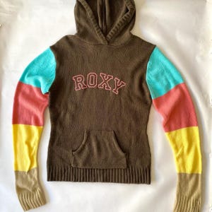 May include: Brown knitted hoodie with a kangaroo pocket and the word "ROXY" in pink lettering. The sleeves are colour-blocked with sections of turquoise, coral, yellow, and tan.