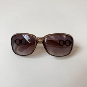 May include: Brown-tinted sunglasses with a square frame. The sunglasses have a gradient lens and decorative oval accents on the temples. The frame is a translucent brown color.