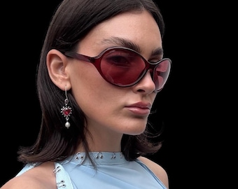 Puede incluir: Una persona lleva gafas de sol de color burdeos con forma ovalada redondeada. También lleva un pendiente de plata con una piedra roja en forma de corazón y una perla colgante. La persona lleva una camiseta azul claro sobre un fondo negro.