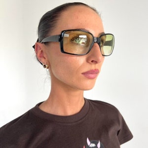 May include: A person wearing black square-shaped sunglasses with yellow tinted lenses. The sunglasses have a thick black frame.