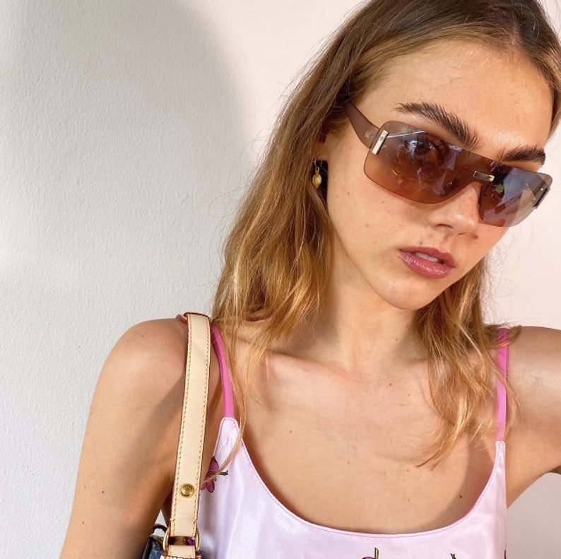 May include: A woman with long blonde hair wearing brown sunglasses and a pink and white floral tank top. She is also wearing a light brown strap of a bag over her shoulder.