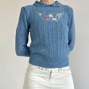 May include: A light blue fuzzy sweater with a white crocheted collar and embroidered flowers in pink, green, and white.
