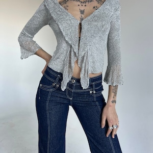 May include: A silver sparkly, open-front cardigan with a ruffled neckline and long sleeves. The cardigan is worn over a pair of dark blue denim jeans.
