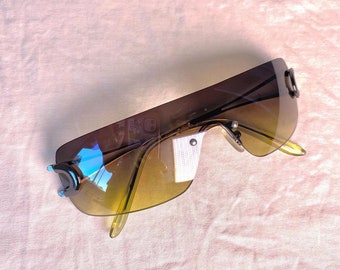 May include: A pair of brown and yellow sunglasses with a unique, curved frame design. The sunglasses have a dark brown frame and lenses with a yellow tint.