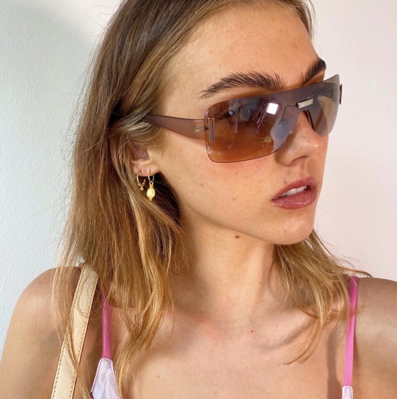 May include: A woman with long blonde hair wearing brown tinted sunglasses with a thin gold chain earring. She is wearing a pink tank top and a light brown strap.