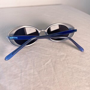 May include: Oval-shaped sunglasses with a translucent silver frame and dark lenses. The temples are a vibrant blue, adding a pop of color. The sunglasses are set against a plain white background.