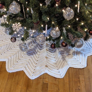 May include: A handmade, off-white crochet Christmas tree skirt with a starburst design. The skirt has a scalloped edge and is secured with wooden buttons. The skirt is under a decorated Christmas tree with ornaments and lights.