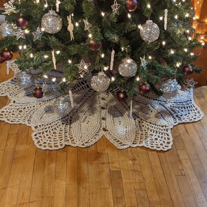 May include: A crocheted cream-colored tree skirt with a scalloped edge, placed under a decorated Christmas tree with silver and burgundy ornaments.