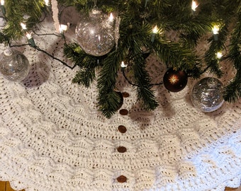 May include: A white crocheted Christmas tree skirt with a scalloped edge and brown buttons down the center. The skirt is decorated with silver and brown ornaments and white lights.