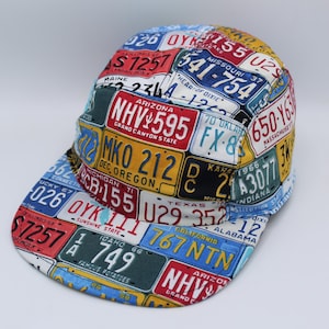 May include: A five-panel baseball cap featuring a colorful all-over print of various vintage American license plates. The design includes plates from states like Arizona, Oregon, Michigan, and California, displaying diverse alphanumeric codes and colors.