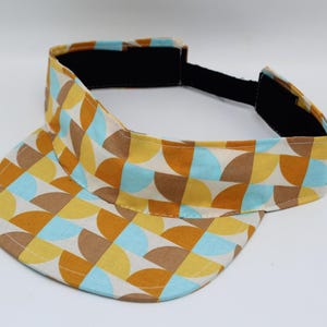 May include: A sun visor featuring a geometric pattern of semicircles in shades of mustard yellow, light blue, and tan on a cream background. The visor includes a black adjustable hook-and-loop strap at the back for a secure fit.
