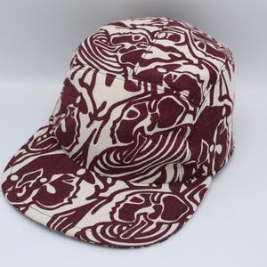 May include: A maroon and cream-colored five-panel cap with a patterned design. The cap features a flat brim and a structured crown. The pattern consists of abstract shapes and lines, creating a unique visual texture.