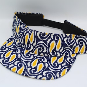 May include: A sun visor featuring a dark blue, white, and yellow paisley pattern. The visor has a curved brim and a black adjustable strap at the back with a hook-and-loop closure. The interior band is lined with black fabric for comfort.