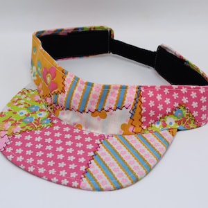Può includere: Una visiera parasole con design patchwork caratterizzata da vari motivi in tessuto, tra cui rosa con fiori bianchi, strisce blu e bianche con fiori rosa e arancione con motivi floreali. La visiera presenta un cinturino nero regolabile.