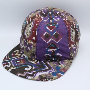 May include: A baseball cap featuring a multi-colored geometric pattern in shades of purple, dark brown, tan, and blue. The cap has a structured crown and a flat brim, both covered in the same intricate, repeating abstract design across the fabric.