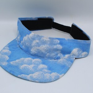 Op de afbeelding: Een lichtblauwe zonneklep met een wit wolkenpatroon op de klep en de hoofdband. De binnenkant van de hoofdband is gevoerd met zwarte stof en heeft een verstelbare zwarte klittenbandsluiting aan de achterkant voor een stevige pasvorm.