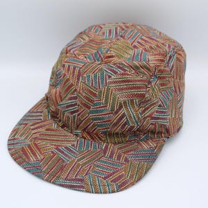 May include: A baseball cap featuring a multi-coloured geometric pattern with repeating chevron-like shapes in shades of red, teal, and gold. The cap has a rounded crown and a flat brim, constructed from a woven fabric with a visible textured weave.