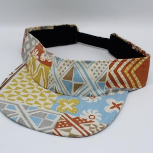 May include: A sun visor featuring a patchwork design with various geometric and floral patterns in shades of light blue, mustard yellow, white, and burnt orange. The visor includes a black adjustable strap at the back and a curved brim.