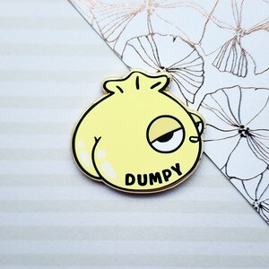 May include: A yellow cartoon dumpling with a single eye and the word "Dumpy" written on it. The dumpling is a gold-colored enamel pin.
