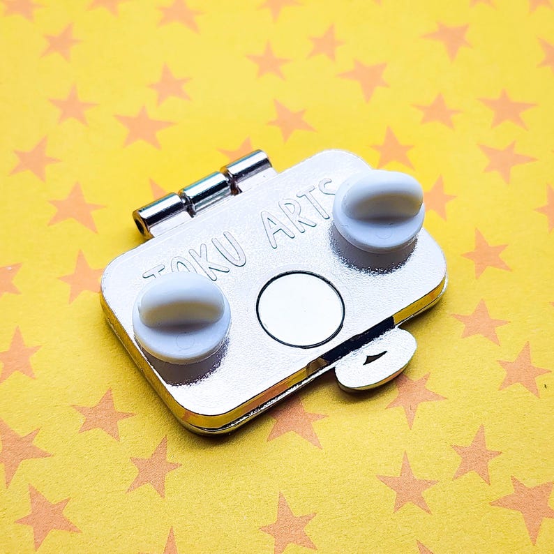 Puede incluir: Parte trasera de un pin de esmalte plateado con dos cierres de goma blancos y un pasador. El pin tiene forma rectangular con esquinas redondeadas y el texto "TOKU ARTS" est&aacute; grabado. Sobre un fondo amarillo con estrellas naranjas.