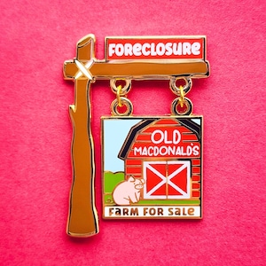 May include: A novelty pin with a farm theme. The pin features a sign that reads "FORECLOSURE" above a sign that reads "OLD MACDONALD'S FARM FOR SALE". The pin has a brown post and gold accents, with a pink pig and red barn.