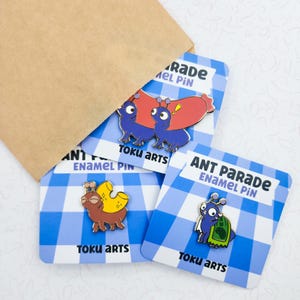 May include: Three enamel pins featuring cartoon ants with the text "ANT PARADE" and "TOKU ARTS". The pins are on blue and white checkered cards, with a brown paper envelope in the background.