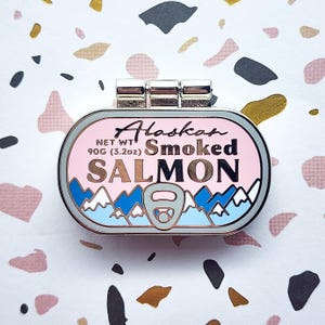 May include: A pin shaped like a can of Alaskan Smoked Salmon. The pin is pink with blue and white mountain graphics. The text on the pin reads "Alaskan Smoked Salmon" and "Net Wt 90g (3.2oz)". The pin has a silver metal top.