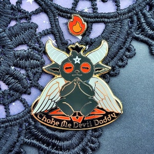 May include: A black and white enamel pin with a cartoon devil with wings and a pentagram on its forehead. The devil is holding its hands together in a praying position. The text "Choke Me Devil Daddy" is written in a gold font around the bottom of the pin.