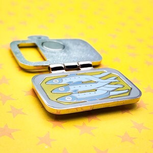Puede incluir: Un pin plateado con bisagras que presenta un dise&ntilde;o de lata de sardinas. La lata est&aacute; abierta, revelando tres peces azules sobre un fondo amarillo. El pin tiene un borde dorado y est&aacute; sobre un fondo amarillo con estrellas.