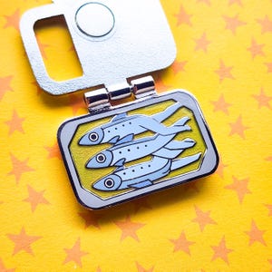 Puede incluir: Un pin de esmalte plateado con tres peces azul claro sobre un fondo amarillo. El pin tiene forma rectangular con borde plateado y cierre magn&eacute;tico. El fondo es una superficie amarilla con formas de estrellas naranjas.