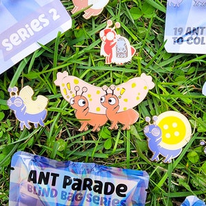 May include: A set of four enamel pins featuring cartoon ants. The pins are colorful and feature different designs, including a snail, a pair of ants carrying a piece of food, and an ant with a large head. The pins are displayed on a green grassy surface with a bag that says "ANT PARADE BLIND BAG SERIES".