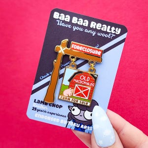 May include: A novelty pin featuring a sign that reads "FORECLOSURE" and "FARM FOR SALE." The pin has a barn design with a pig and the words "OLD MACDONALD'S." The pin is on a card that says "Baa Baa Realty" and "Lamb Chop Realtor."