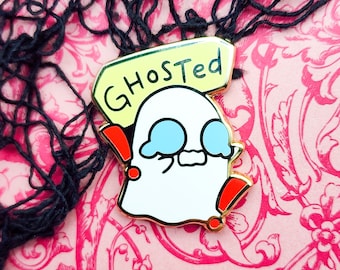 Puede incluir: Un pin de esmalte con forma de fantasma blanco con la palabra "GHOSTed" en un letrero amarillo. El fantasma tiene ojos azules y signos de exclamación rojos a los lados. El pin tiene un borde dorado y está sobre un fondo floral rosa.