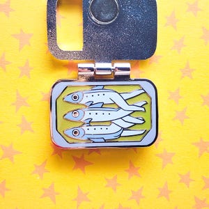 Puede incluir: Una llave plateada con un pin rectangular de esmalte que representa tres peces azul claro estilizados sobre un fondo verde. El pin tiene un borde plateado, sobre un fondo amarillo con estrellas naranjas.